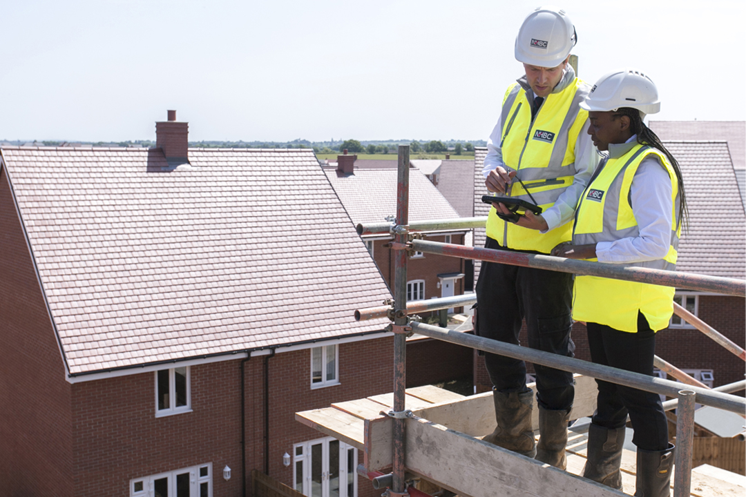 two people in site safety clothing and hard hats stand on scaffolding and look at plans