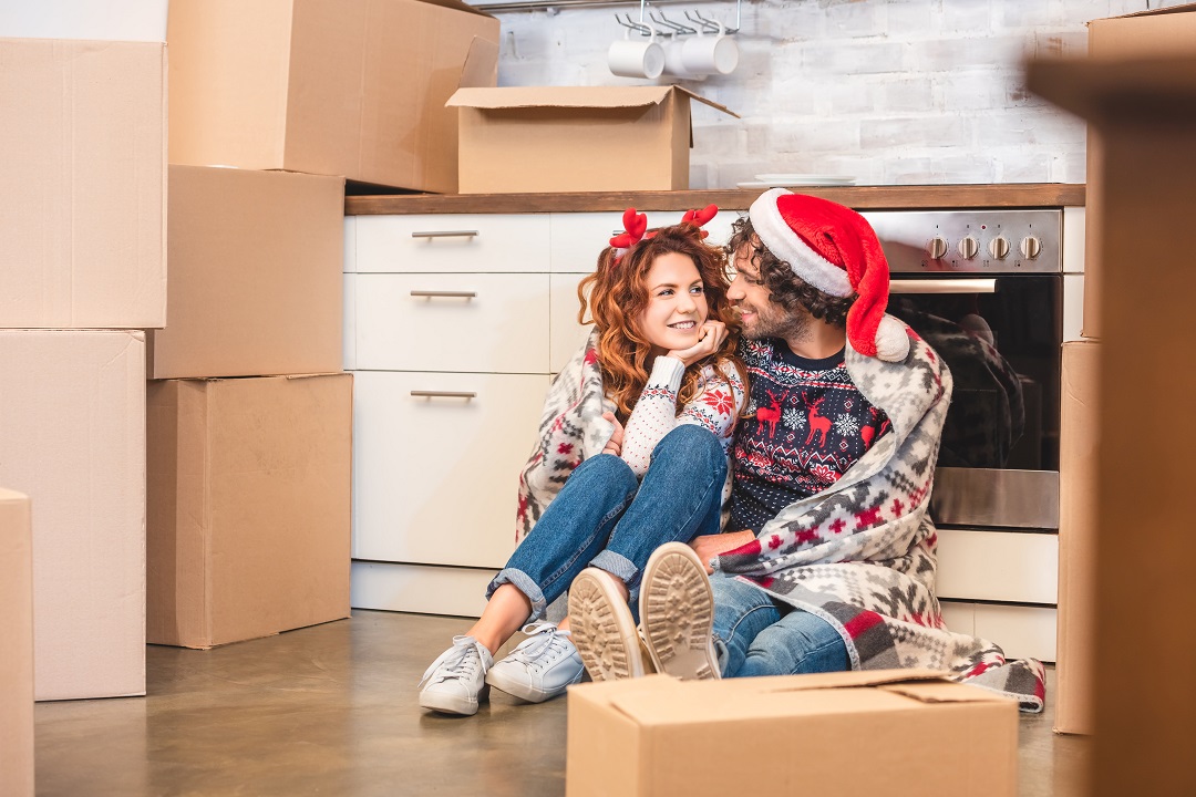 a couple wearing christmas clothing sat in their kitchen surrounded by boxes