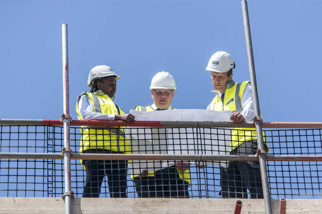 three people in site safety clothing, stood on scaffolding on a sunny day
