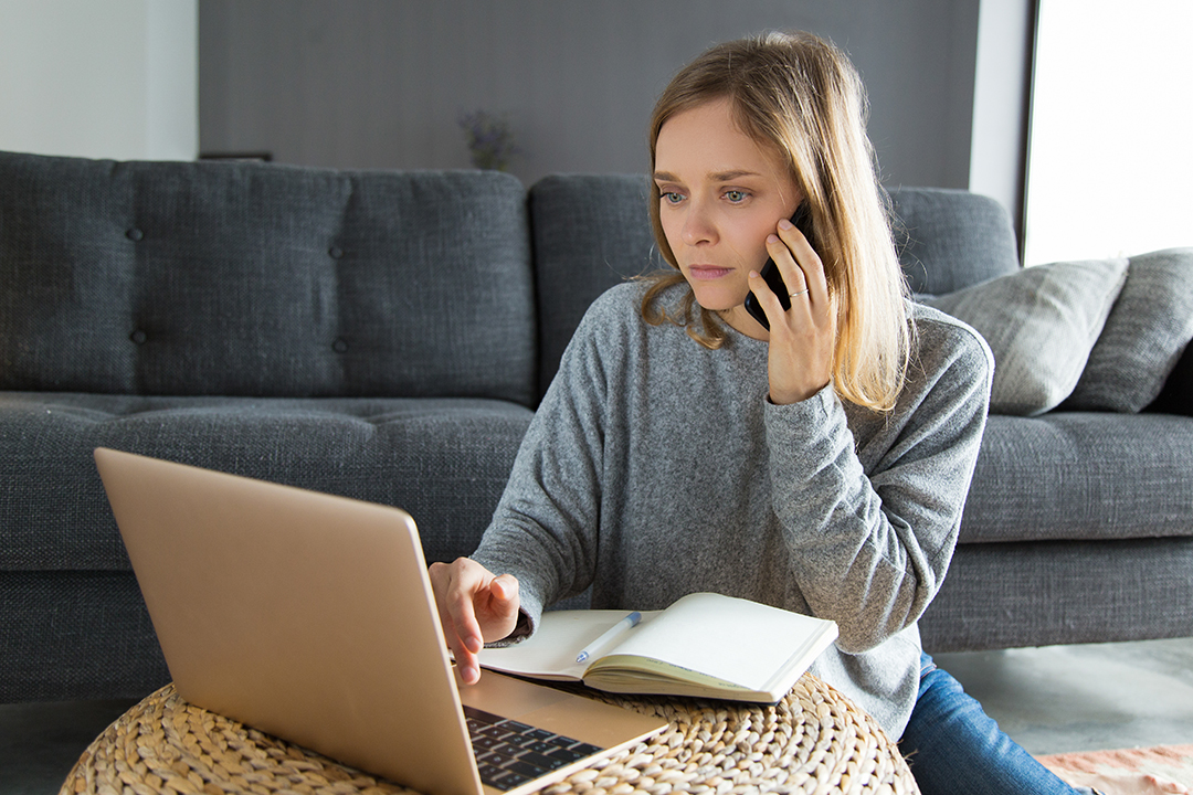 a woman in a grey jumper on the phone while looking at a laptop