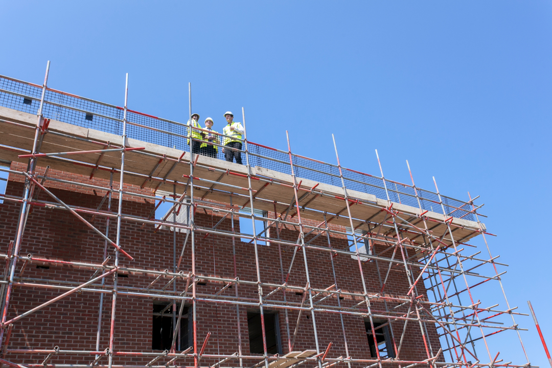 three people in site safety clothing and hard hats stand on some scaffolding on a sunny day