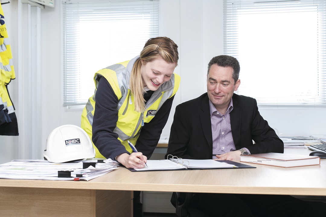 two people in an office, one wearing site safety clothing, signing a document