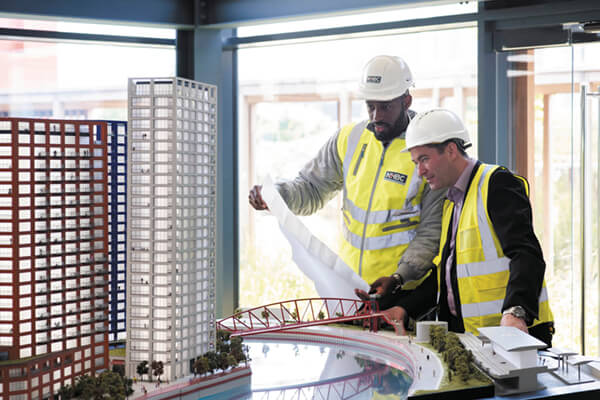 two people in site safety clothing and hard hats looking at plans and a scale model of a development