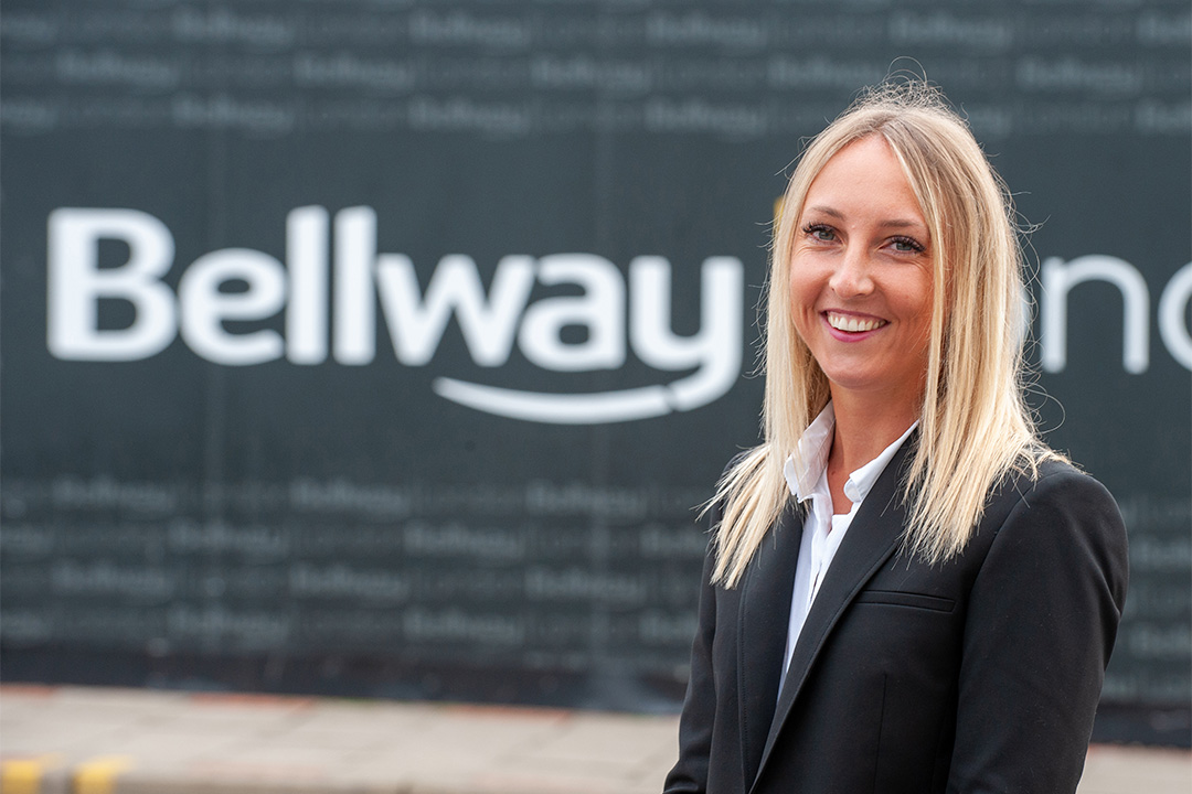 a blonde woman ia blazer smiling at the camera in front of a bellway sign
