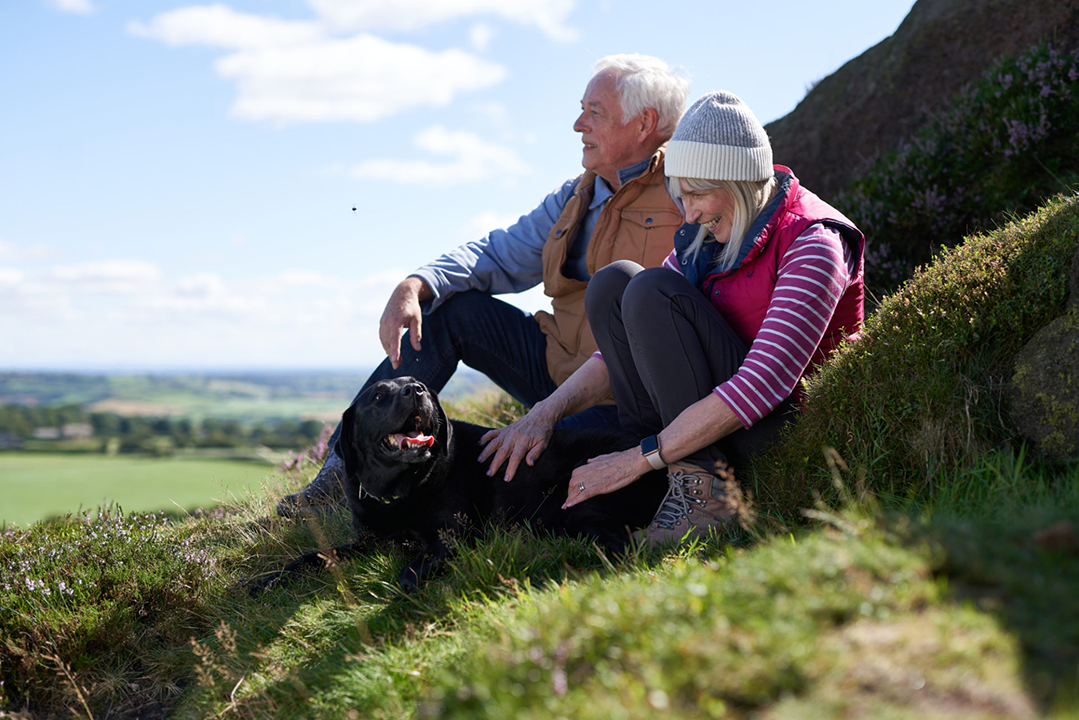 an older couple and their black dog sitting on the hillside