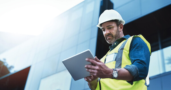 a man in site safety clothing and a hard hat looking at a tablet