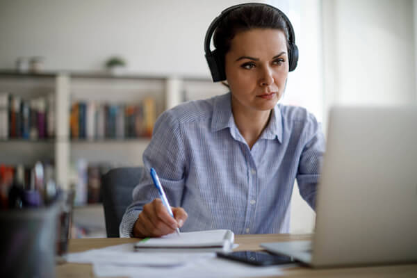 a woman wearing headphones looking at a laptop and taking notes